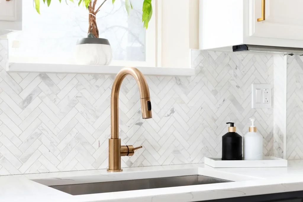 Sink detail shot in a luxury kitchen with herringbone backsplash tiles. white marble countertop, and gold faucet.